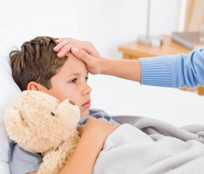Ein kleiner Junge liegt krank mit Teddy im Bett und die Mutter hat die Hand auf seiner Stirn, um Temperatur zu messen.